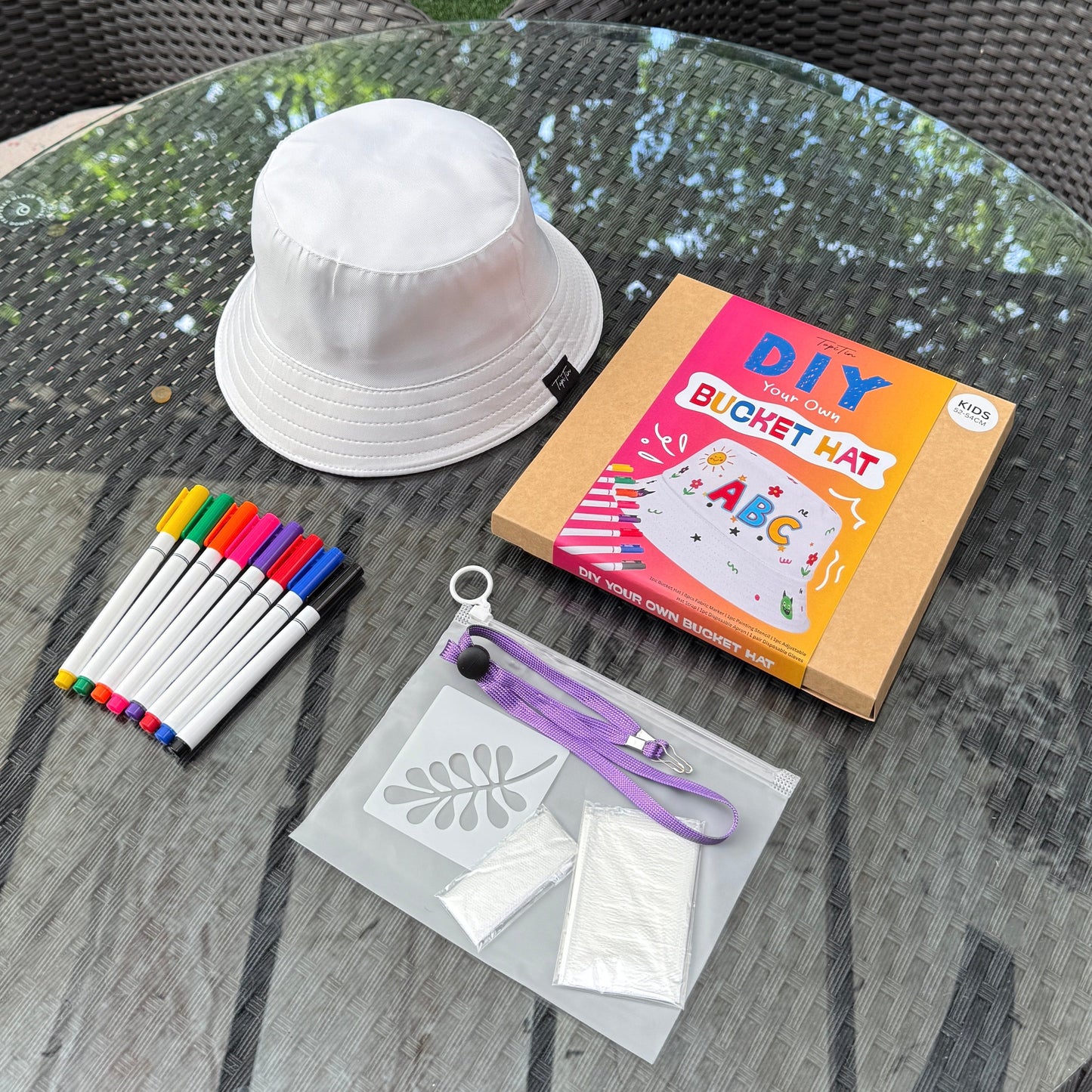 Round outdoor table with DIY bucket hat kit, bucket hat, markers, and stencils on a green grass background.